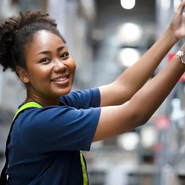 Eine lächelnde schwarze Frau in einem blauen Hemd und mit einer Armbanduhr greift nach oben, um Kisten auf einem hohen Regal in einem hell erleuchteten Lagerhaus zu ordnen. Sie ist wahrscheinlich eine Mitarbeiterin von Über Hermes Fulfilment, die für eine effiziente Auftragsabwicklung sorgt.